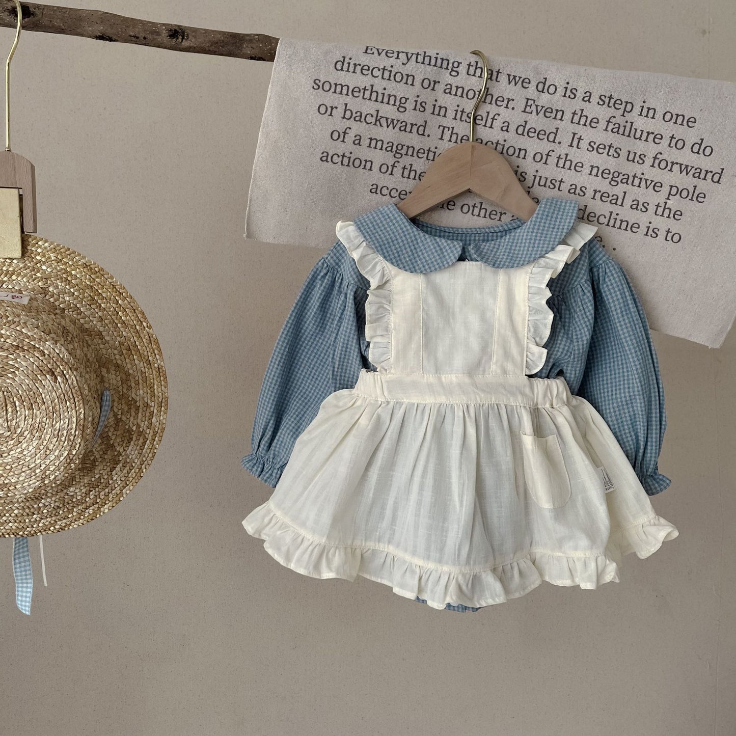 Children's outfit with white dress and blue shirt hanging on a wooden hanger against a beige wall.