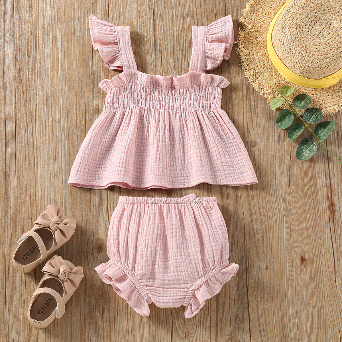 Pink baby outfit with top and bloomers on a wooden surface, accompanied by shoes and a hat.