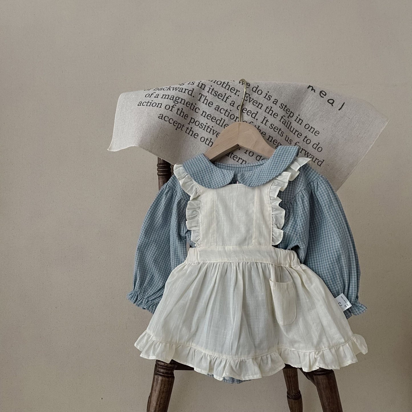 Children's outfit with blue checkered blouse and white apron on a wooden hanger against a beige background.