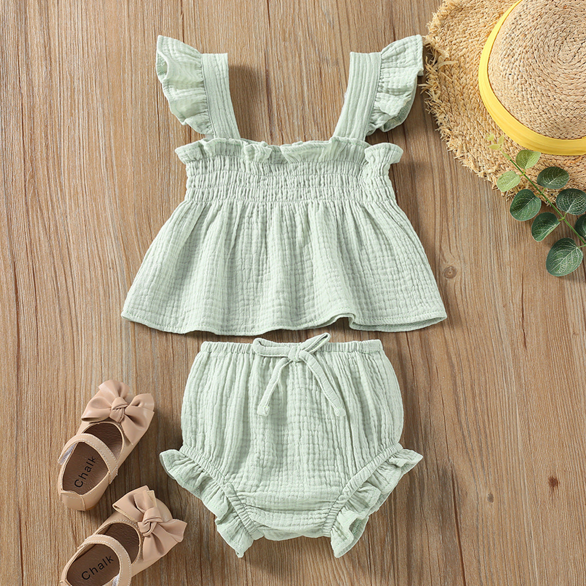 Mint green baby outfit with top and shorts on a wooden surface, accompanied by shoes and a hat.
