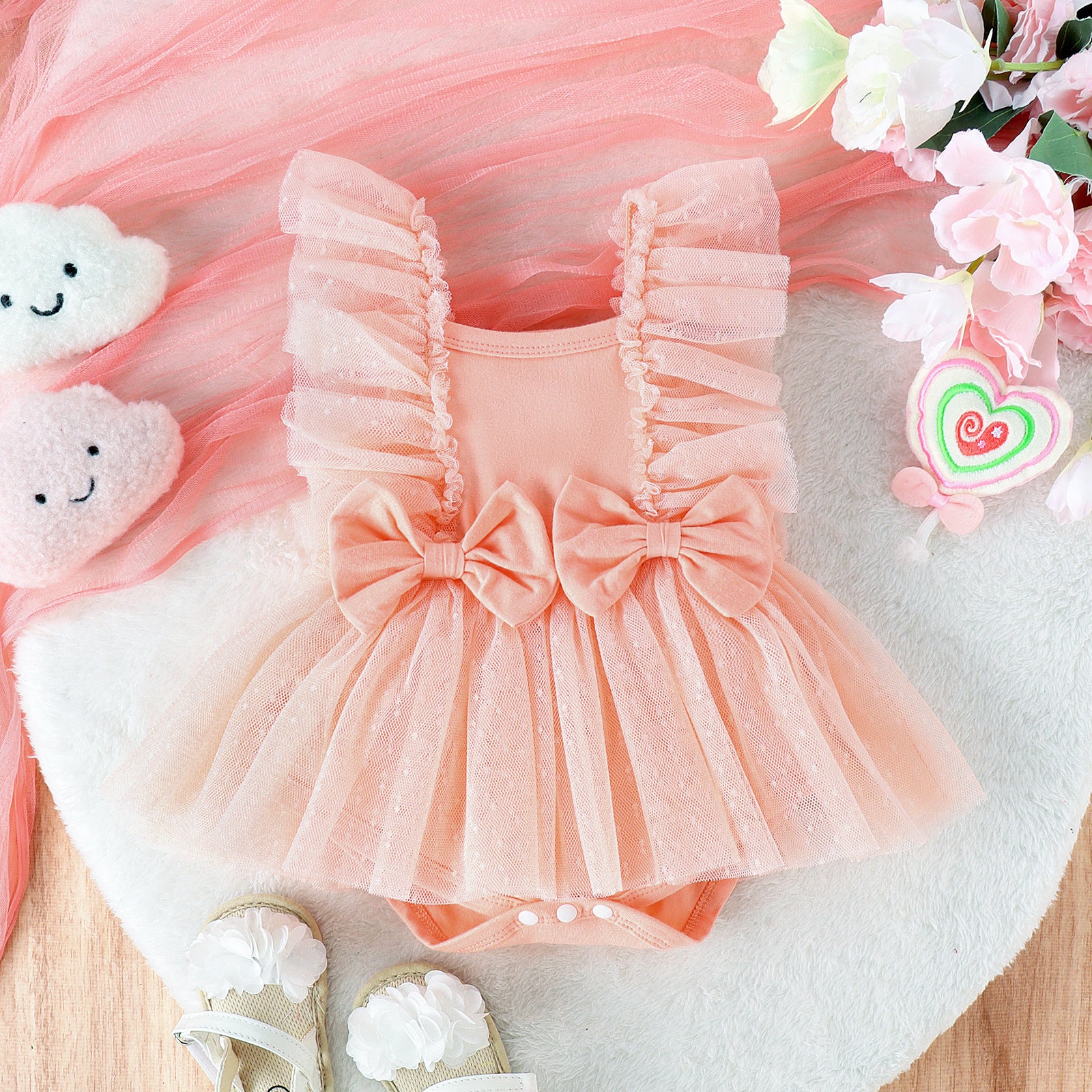 Pink baby romper with ruffles and a bow, surrounded by flowers and toys on a light surface.