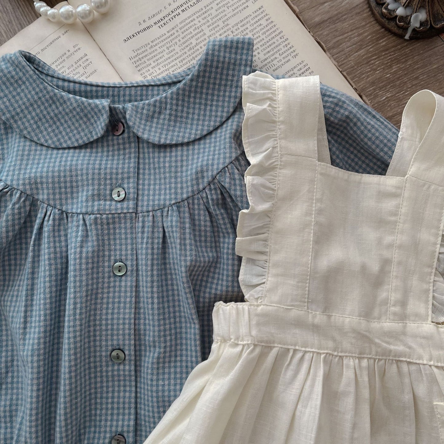 Two children's dresses, one blue checkered and one beige, on a wooden surface with a book underneath.