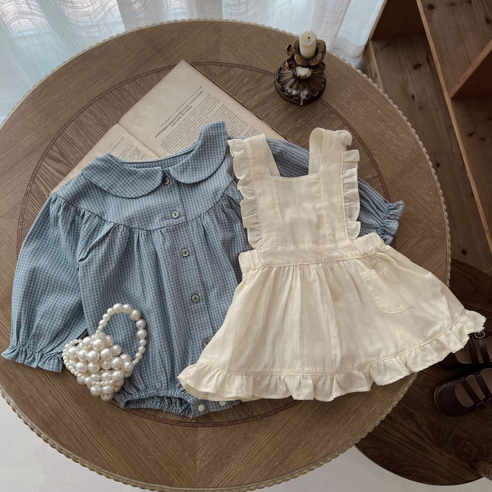 Children's blue checkered shirt and white dress on a wooden table with a pearl basket.
