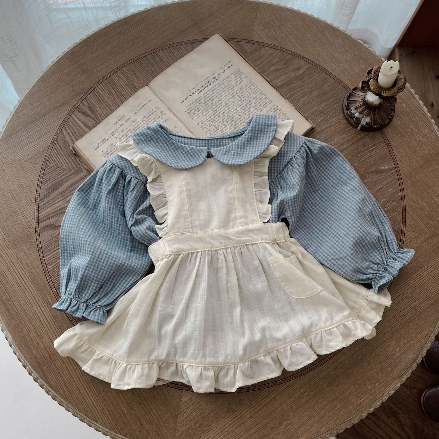 Children's outfit with white dress and blue checkered shirt on a wooden table.